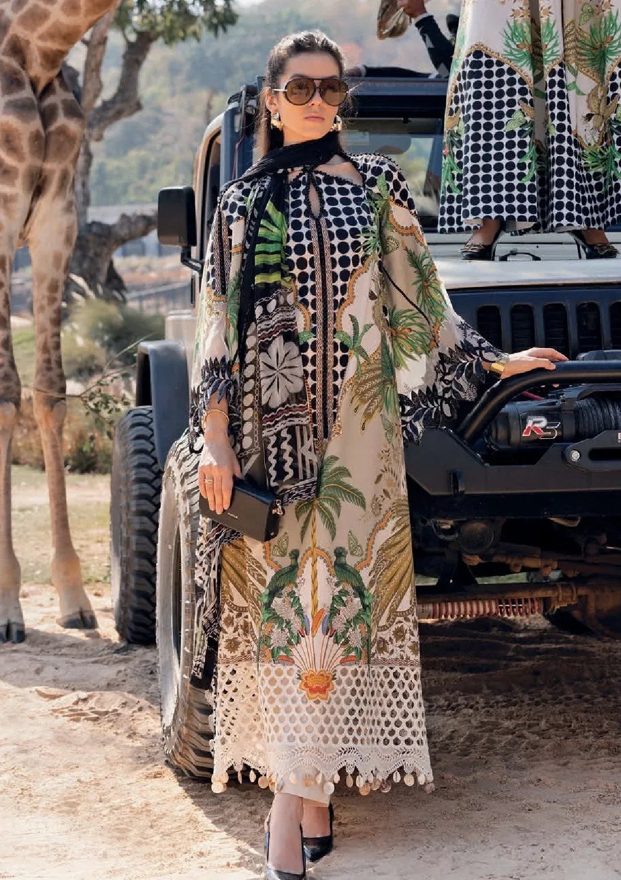 A woman wearing a long beige and black patterned kaftan dress with green botanical prints stands outdoors next to a black off-road vehicle, with a giraffe visible on the left.