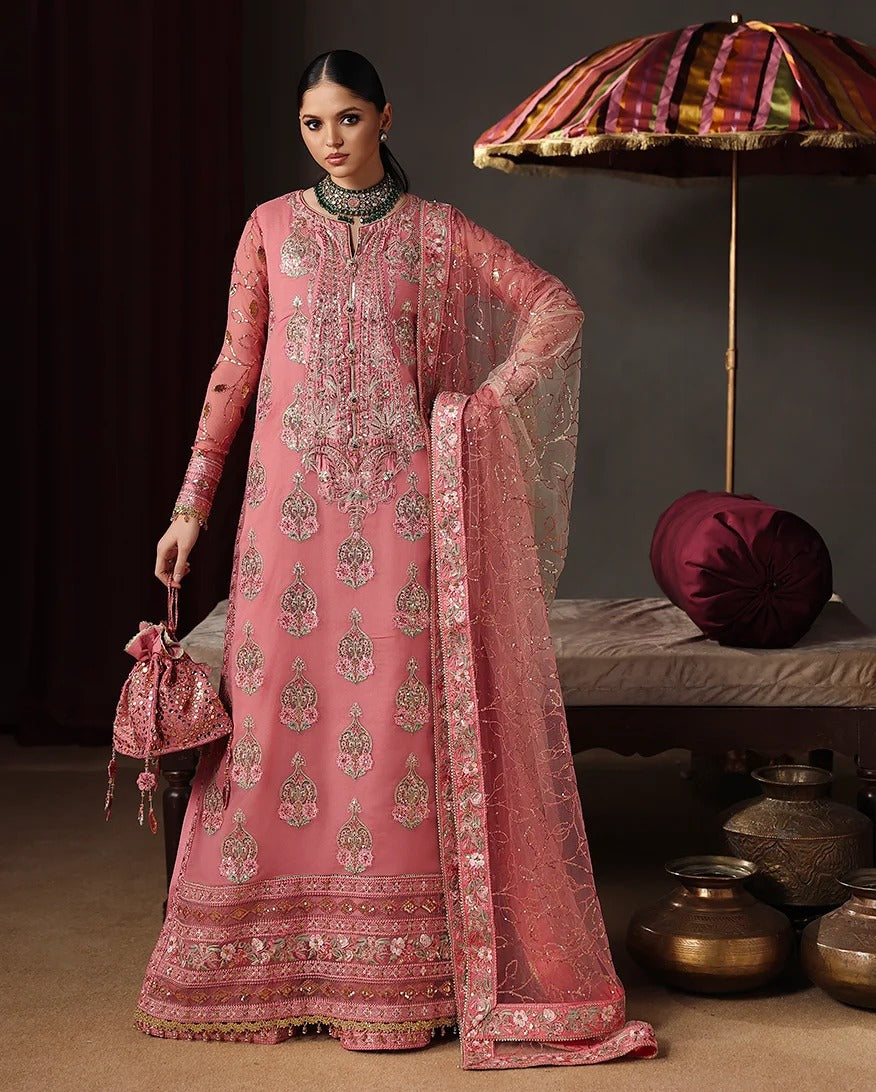 A woman models a traditional pink embroidered Indian outfit with a matching dupatta, standing in a styled studio setting with a decorative umbrella, cushions, and metal pots in the background.