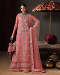 A woman models a traditional pink embroidered Indian outfit with a matching dupatta, standing in a styled studio setting with a decorative umbrella, cushions, and metal pots in the background.