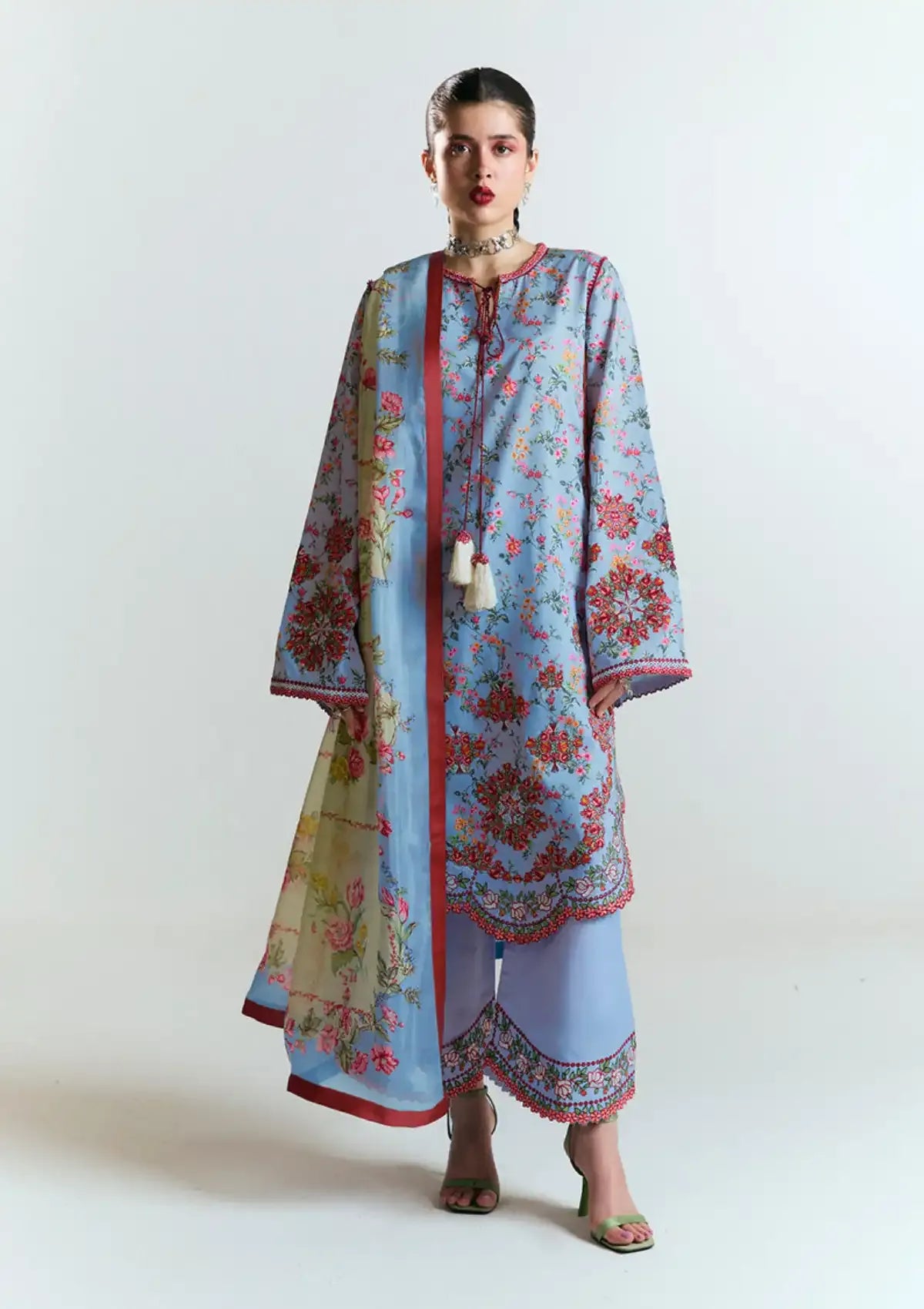 A model wearing a light blue three-piece salwar kameez suit with red and floral patterns, a matching dupatta, and light blue trousers, photographed against a plain white studio background.