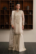 A woman models a traditional South Asian outfit consisting of a cream-colored embroidered kurta, matching wide-leg trousers, and a sheer dupatta, set against a dark wood shelving unit.