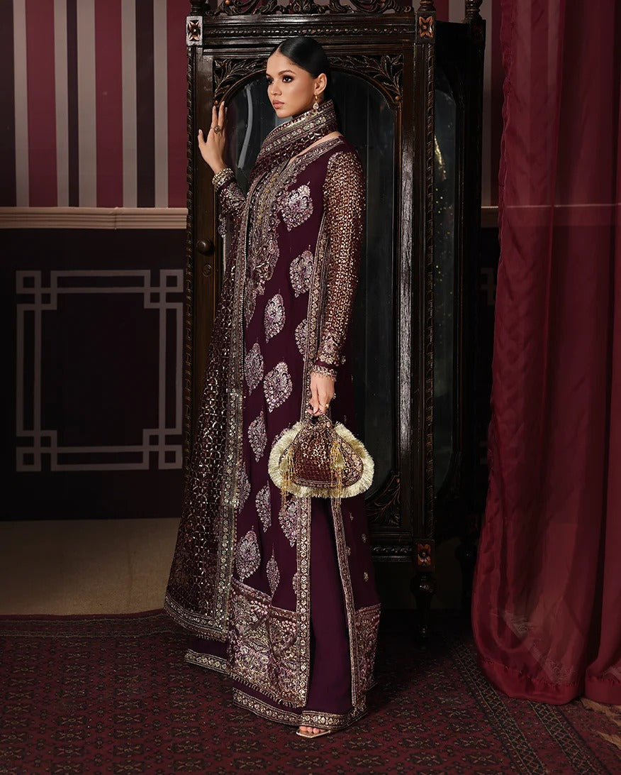 A woman models an ornate deep maroon and gold embroidered traditional outfit, standing in front of a dark carved cabinet in an elegant room with red curtains and patterned rugs.