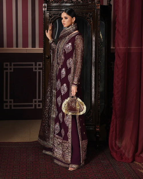 A woman models an ornate deep maroon and gold embroidered traditional outfit, standing in front of a dark carved cabinet in an elegant room with red curtains and patterned rugs.