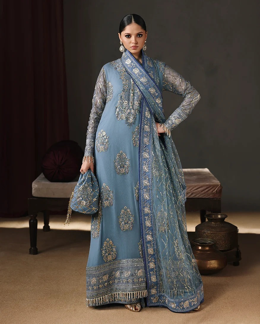 A woman models a traditional blue and gold embroidered Indian outfit with a matching dupatta and clutch, posed in front of a dark studio backdrop with decorative furniture.