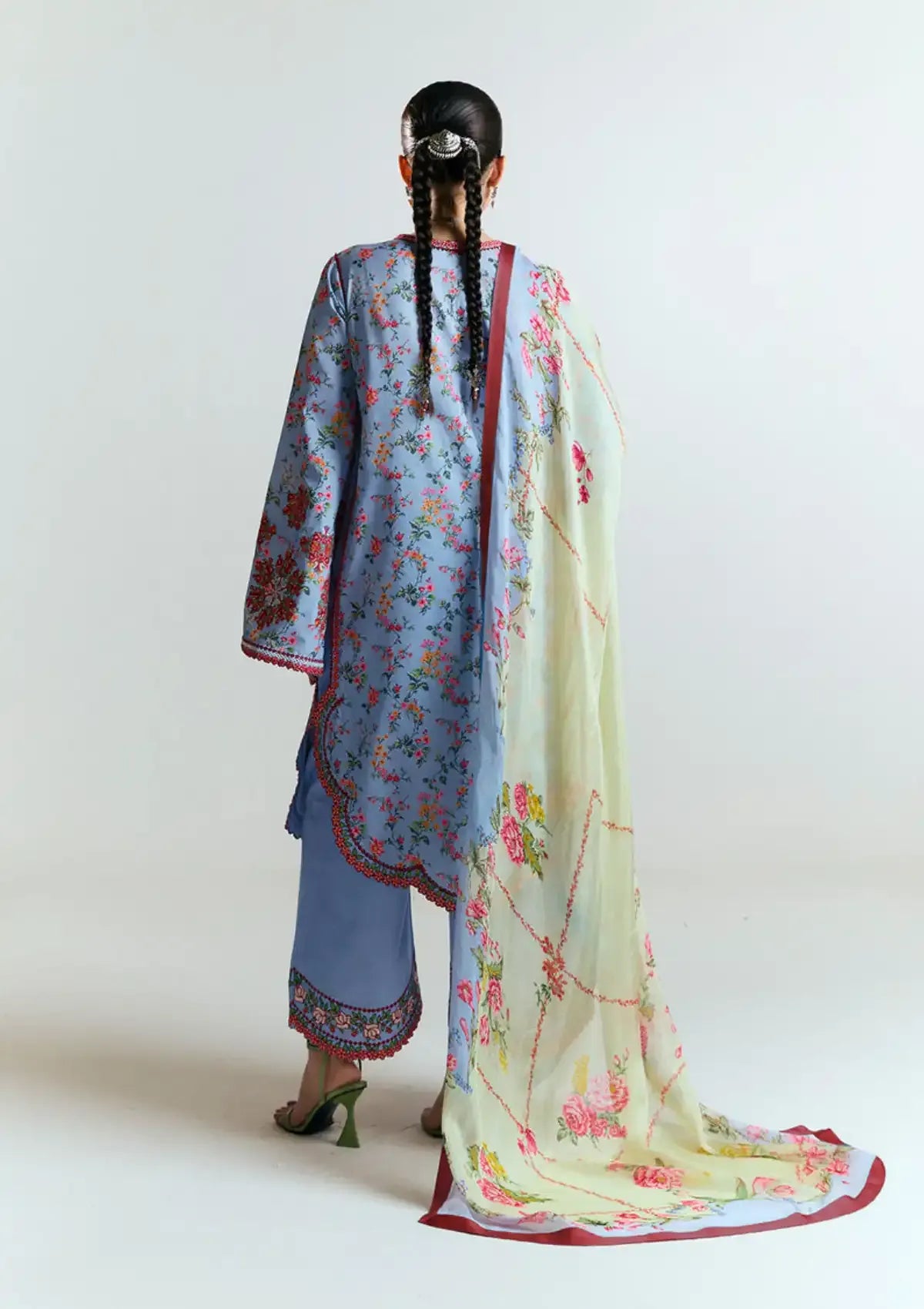 A woman shown from the back wearing a light blue floral traditional outfit with a long cream dupatta featuring pink floral patterns and red borders, set against a plain light gray studio background.
