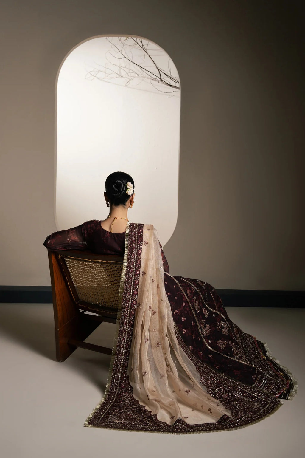 A woman seated on a wooden chair, viewed from behind, wearing an ornate dark plum and beige embroidered traditional outfit with a sheer patterned drape. She is posed in front of a large arched window-like opening against a warm gray wall.