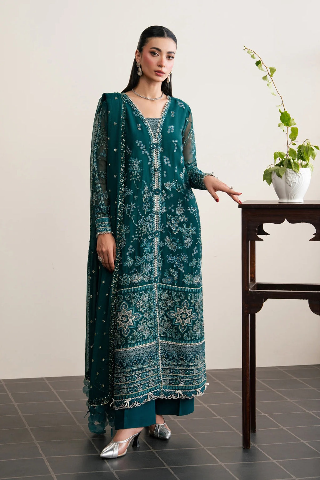 A woman models a teal green embroidered traditional kurta set with matching pants and silver heels, standing next to a dark wooden table with a potted plant against a neutral beige wall.