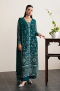 A woman models a teal green embroidered traditional kurta set with matching pants and silver heels, standing next to a dark wooden table with a potted plant against a neutral beige wall.