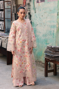 A woman stands in a rustic shop setting wearing a long, light pink embroidered kurta with a floral print and matching wide-leg pants. The kurta features intricate lace and embroidery, and the background shows a weathered teal wall and wooden tables.