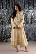 A woman models a traditional long kurta set in a pale beige or cream color with intricate green and orange floral embroidery. She is standing in front of a silver tinsel curtain backdrop.