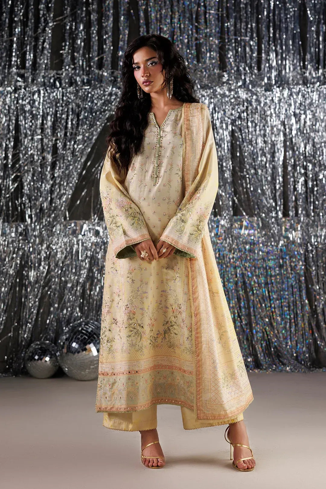 A woman models a traditional long kurta set in a pale beige or cream color with intricate green and orange floral embroidery. She is standing in front of a silver tinsel curtain backdrop.