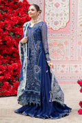 A woman models a traditional navy blue Indian ethnic outfit with intricate silver embroidery, a matching dupatta, and a flared skirt, posed in front of a decorative wall with red flowers.