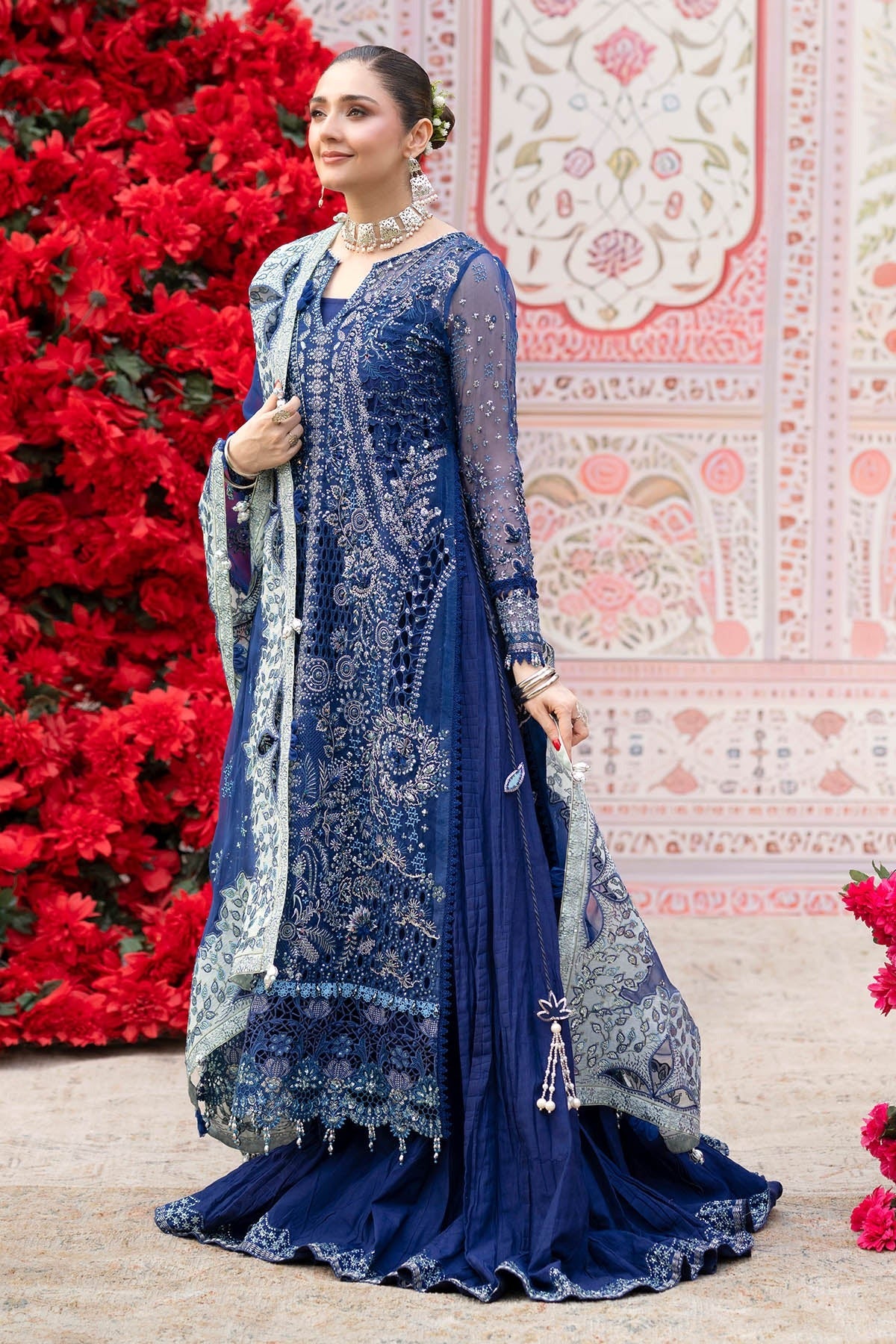A woman models a traditional navy blue Indian ethnic outfit with intricate silver embroidery, a matching dupatta, and a flared skirt, posed in front of a decorative wall with red flowers.