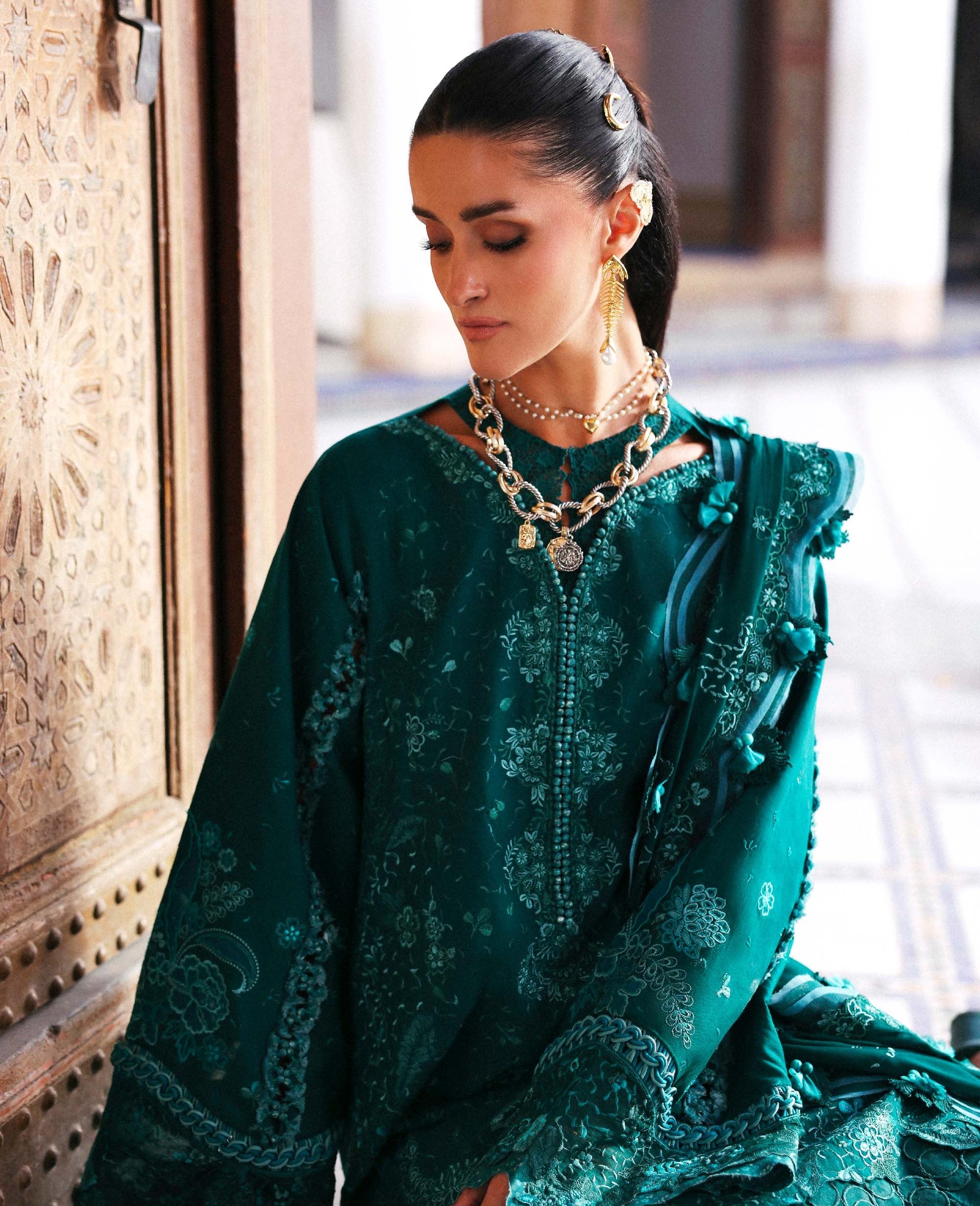 Woman wearing a green embroidered traditional outfit with jewelry, standing against a blurred background.