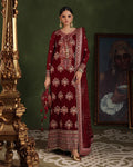 A woman models a deep maroon red traditional embroidered kurta set with a matching dupatta, standing in a dark green room with an ornate gold pillar and a framed painting in the background.