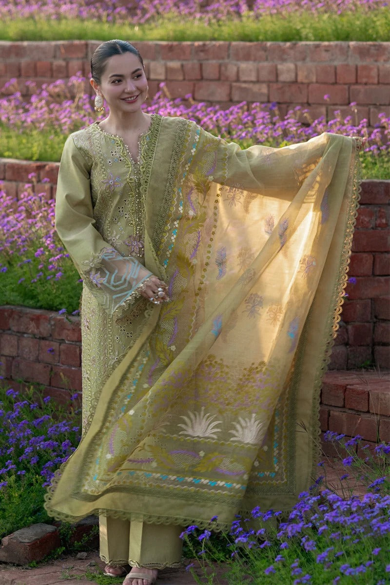 Woman in a green traditional outfit with a matching dupatta, standing in a garden with purple flowers and a brick wall.