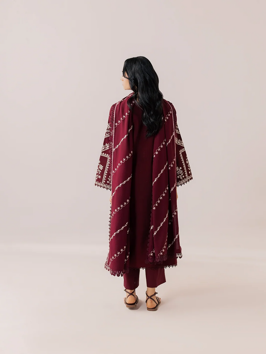 A woman is shown from behind wearing a deep maroon traditional outfit with a matching embroidered shawl featuring white geometric patterns, set against a plain light beige studio background.