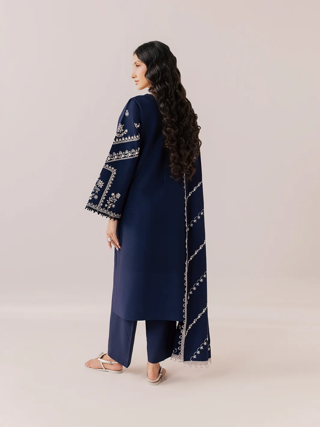 A woman modeling a navy blue traditional outfit with white embroidery, shown from a side-back angle against a solid light beige studio background.