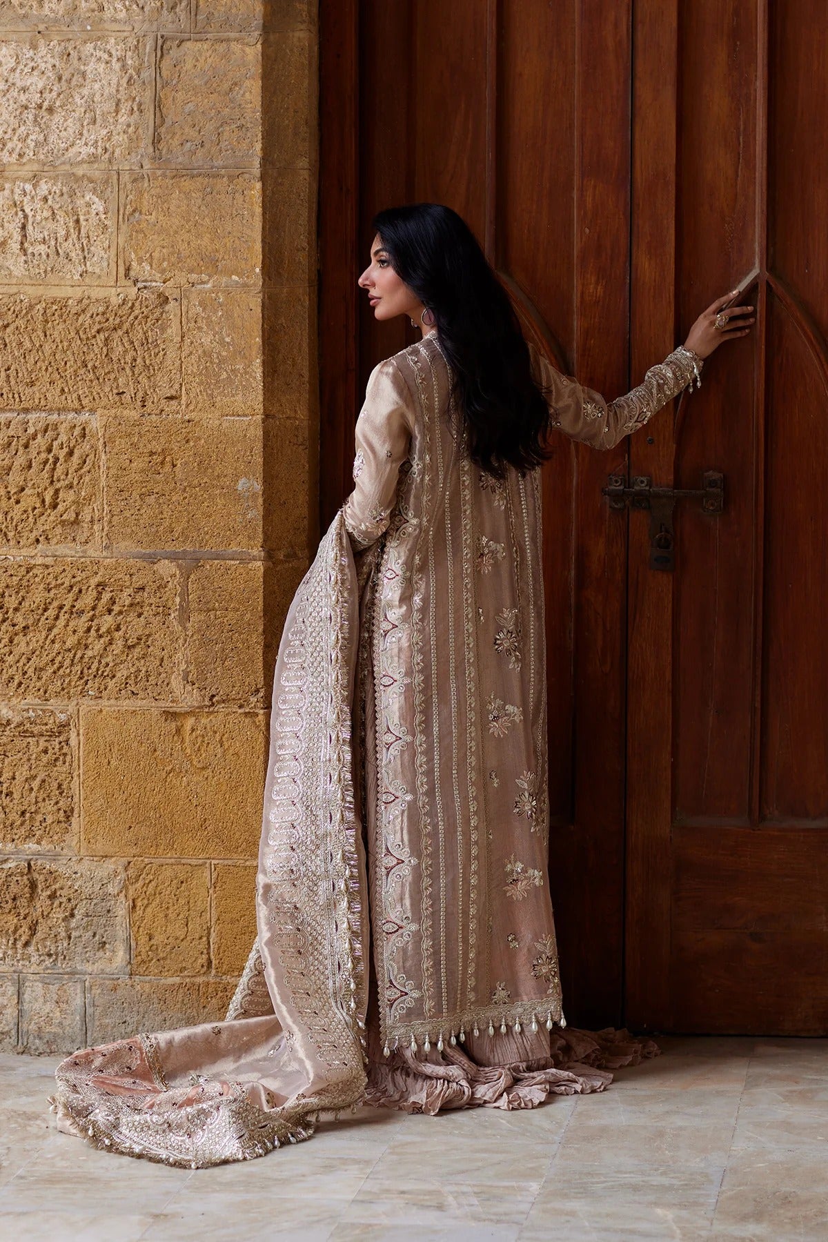 A woman in a traditional beige embroidered outfit with a matching dupatta stands beside a large wooden door, touching the door with one hand. The setting features a textured stone wall on the left and a polished wooden door on the right.