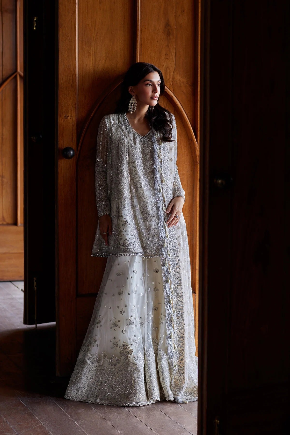 A woman wearing a traditional white embroidered kurta set with a matching skirt, standing in a doorway with dark wood paneling.