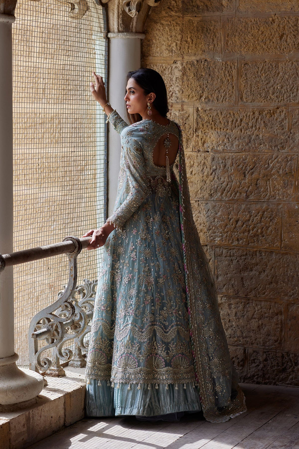 A woman wearing a light blue heavily embroidered traditional gown stands by a stone wall and window in a historic building, holding onto a metal railing.