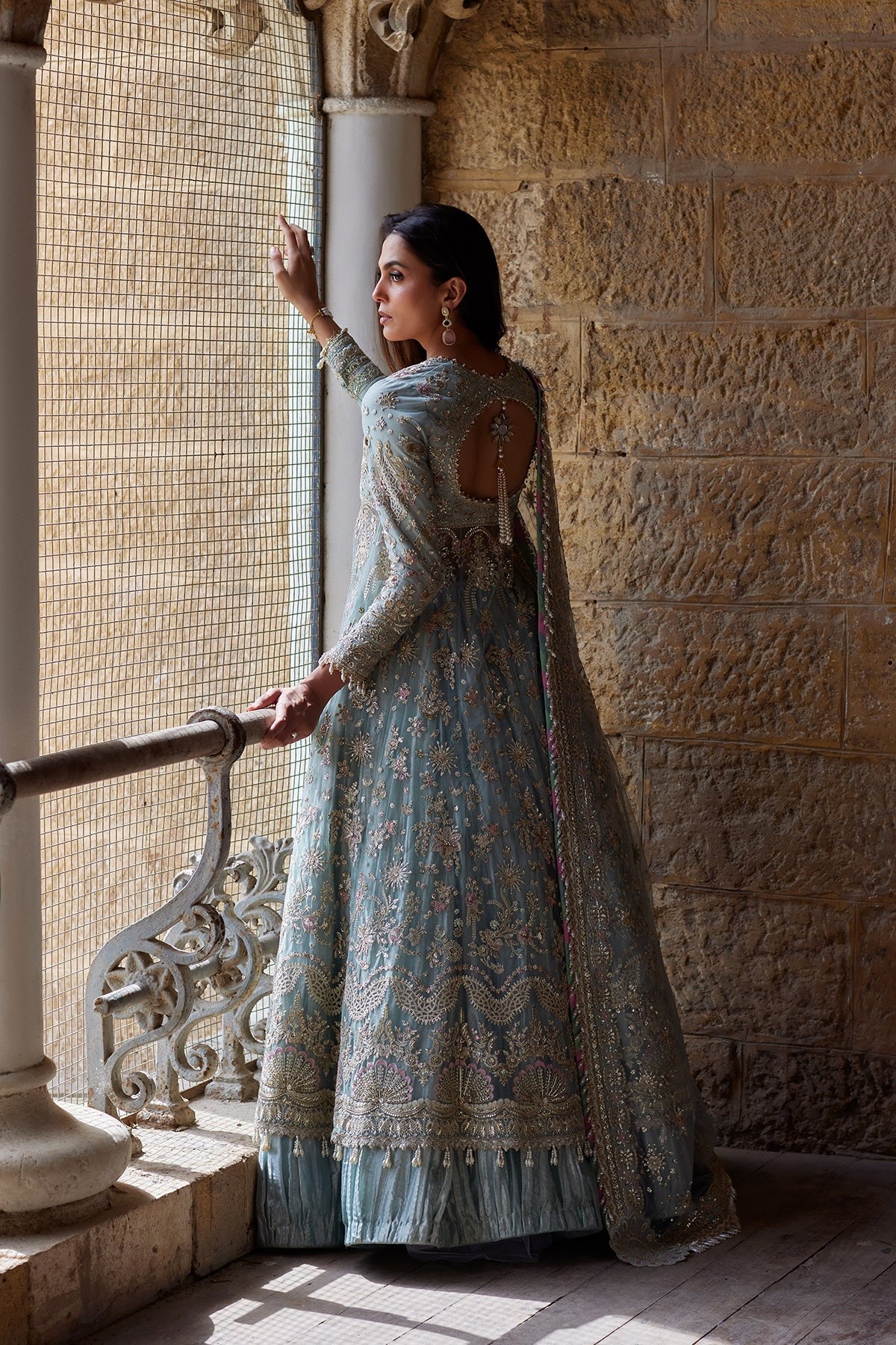 A woman wearing a light blue heavily embroidered traditional gown stands by a stone wall and window in a historic building, holding onto a metal railing.