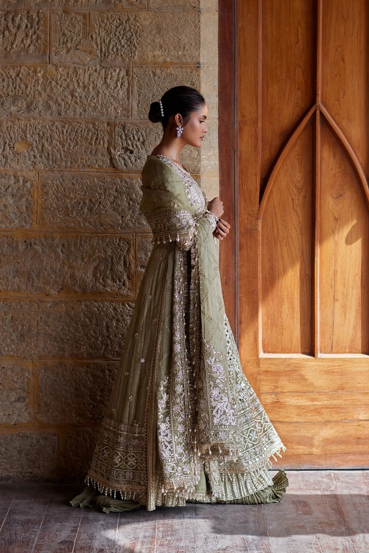 A woman in profile wearing an ornate olive green embroidered traditional gown with a matching draped scarf, standing beside a large wooden door against a stone wall.