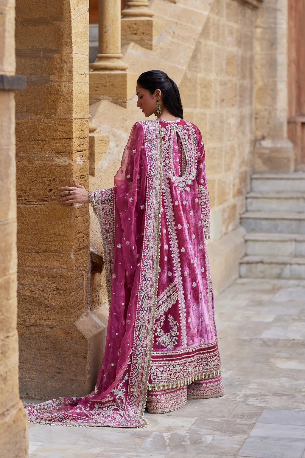 A woman wearing a traditional pink embroidered kurta set with a matching dupatta, standing in a stone corridor.