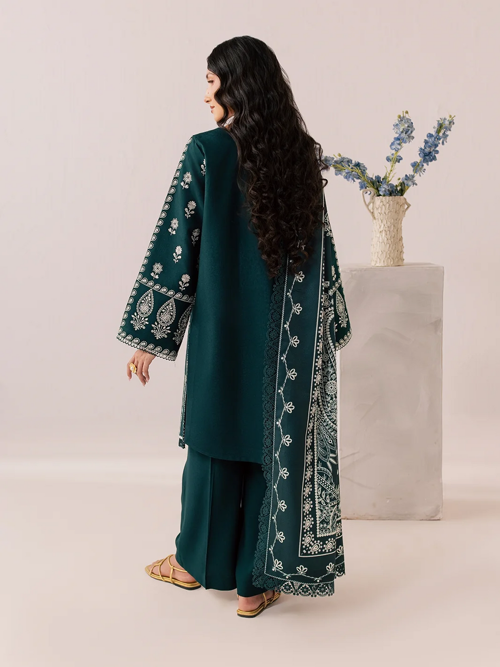 A woman modeling a dark teal three-piece traditional outfit with white embroidery, shown from the back. The set includes a long kurta, wide-leg pants, and a matching dupatta draped over her shoulder.