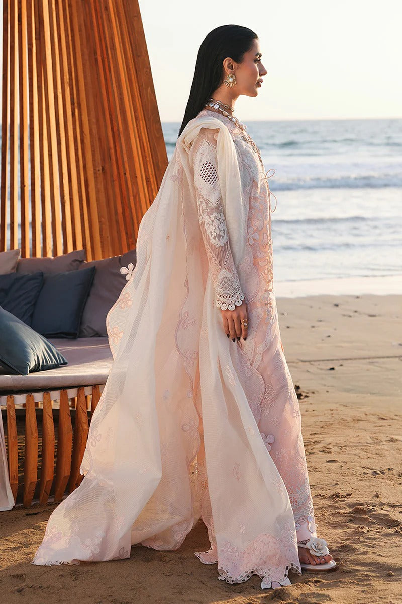 Woman in a light pink traditional outfit standing on a beach with ocean and wooden structure in the background.