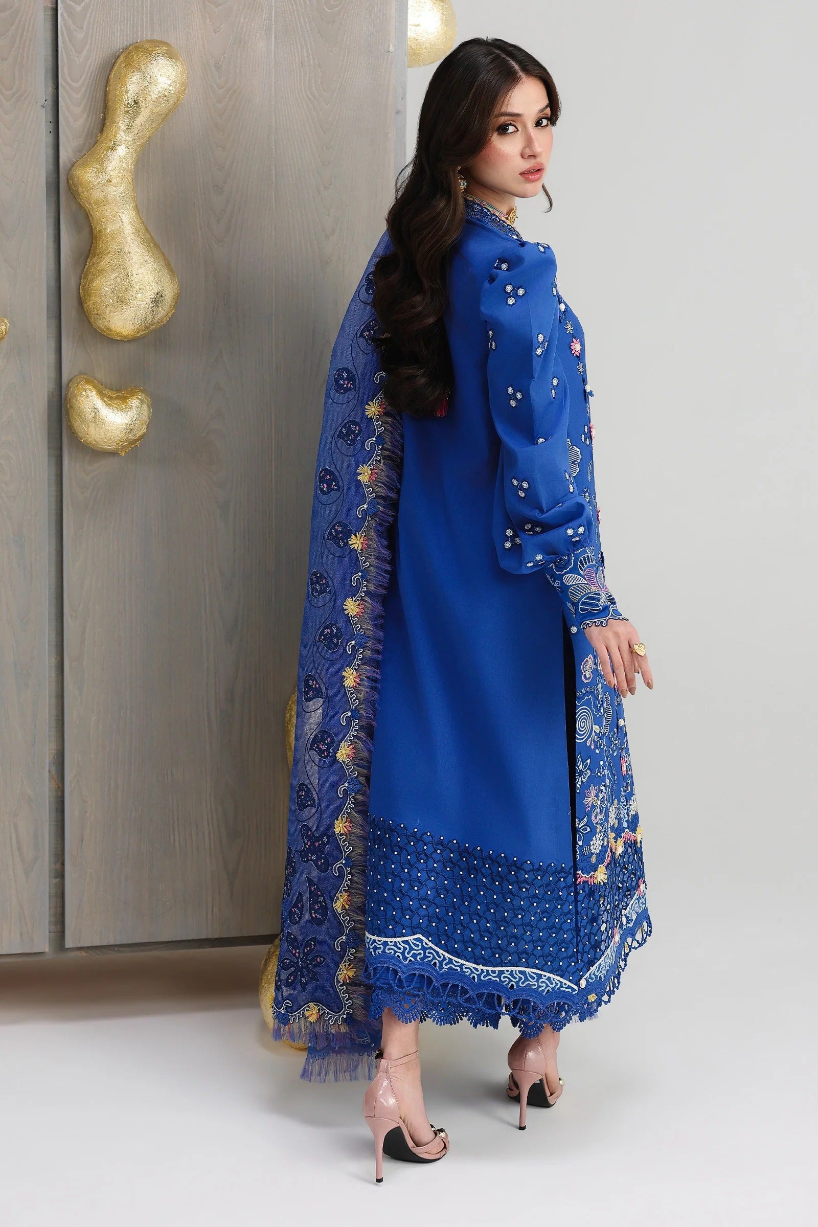 Woman in a blue traditional outfit with gold embroidery standing against a neutral background