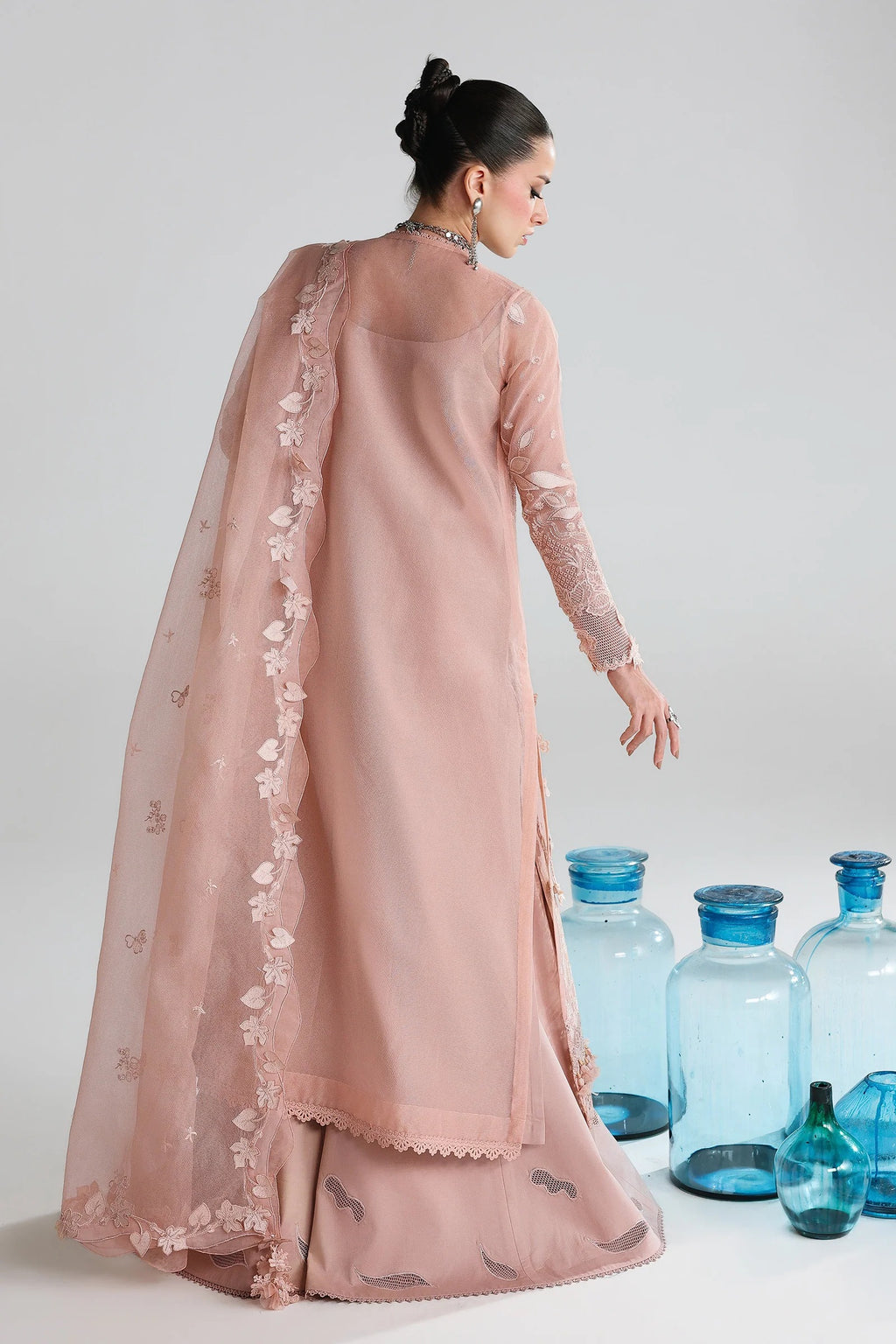 Woman in a pink traditional outfit with a sheer dupatta, standing against a white background with blue bottles.