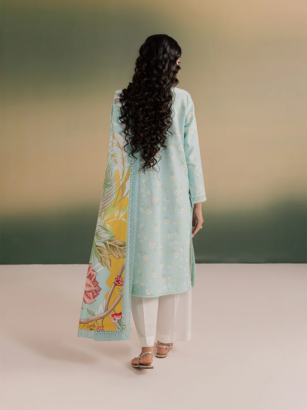 A woman shown from the back wearing a light blue embroidered kurta with a matching floral printed dupatta and white pants, standing against a soft gradient studio background.