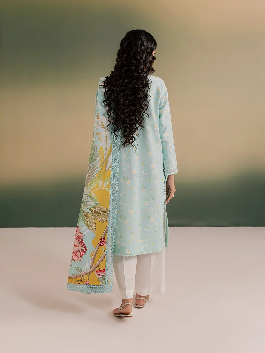 A woman shown from the back wearing a light blue embroidered kurta with a matching floral printed dupatta and white pants, standing against a soft gradient studio background.