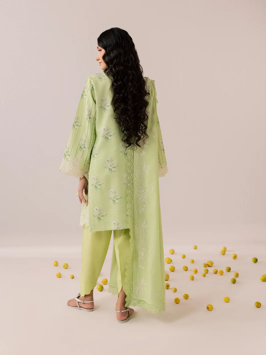 A woman stands with her back to the camera wearing a light green embroidered kurta and matching pants. The outfit features delicate floral embroidery and lace trim.