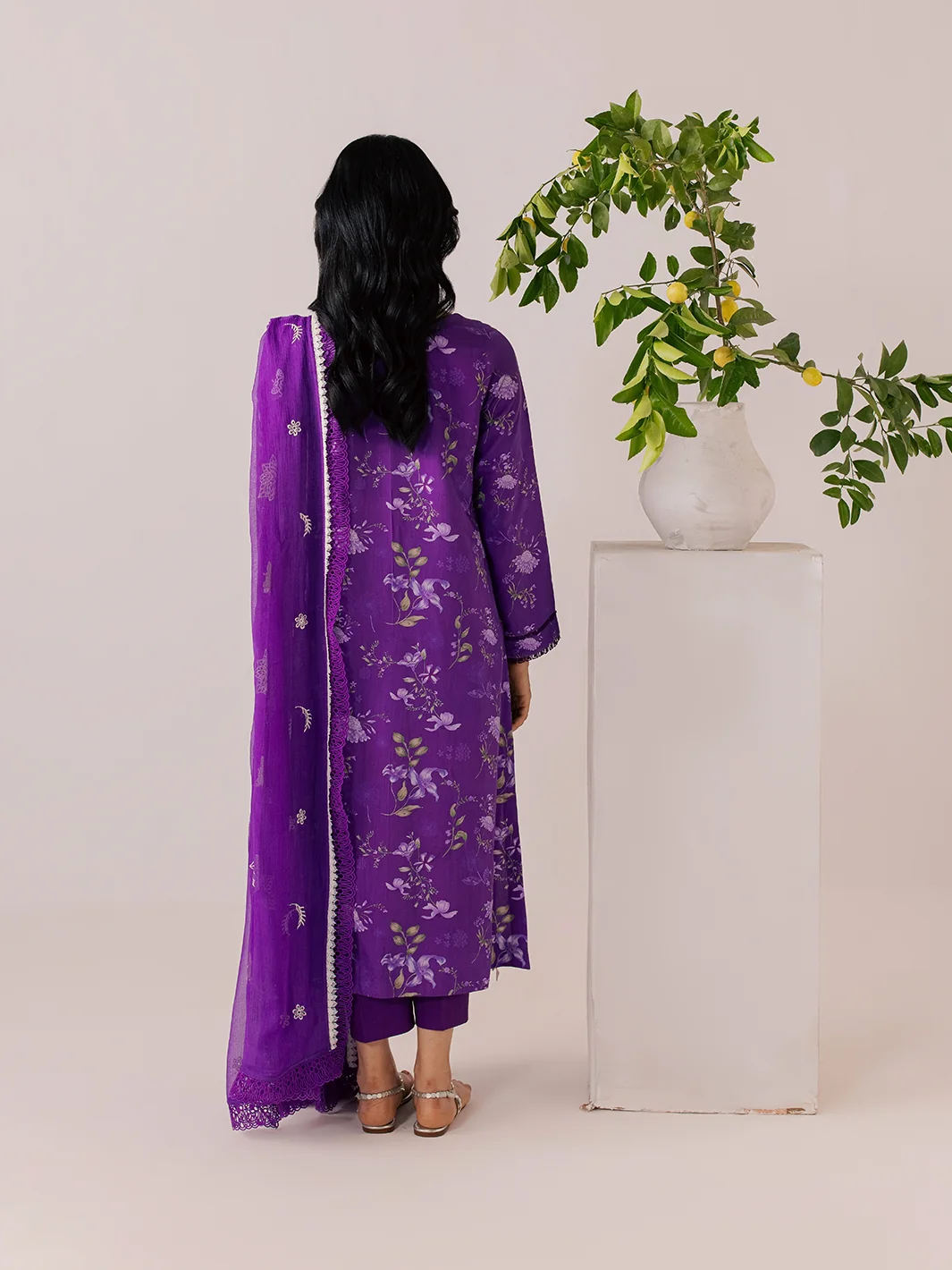 A woman stands with her back to the camera wearing a purple floral traditional outfit with a matching sheer dupatta. She is posed next to a white pedestal holding a potted green plant, against a solid light beige studio background.