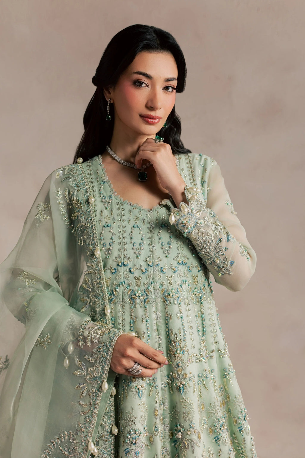 A woman modeling a light sage green embroidered traditional outfit with sheer sleeves and a matching dupatta, set against a soft beige studio background.