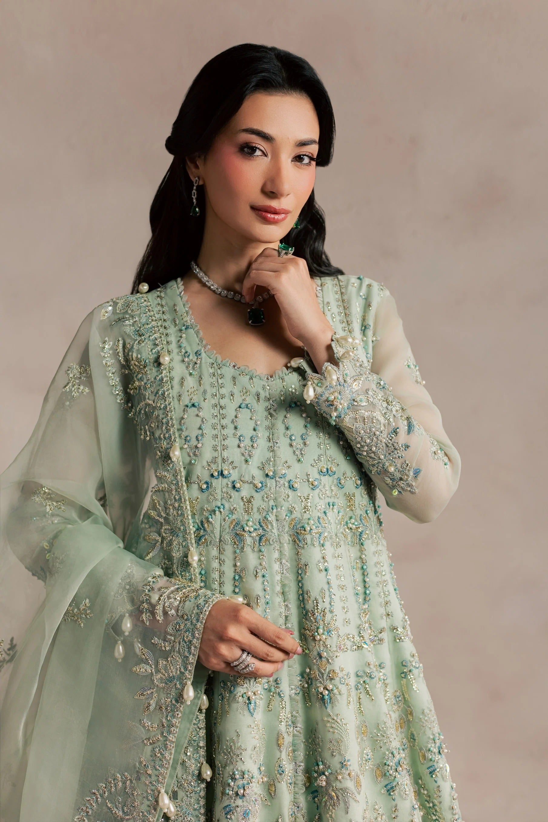 A woman modeling a light sage green embroidered traditional outfit with sheer sleeves and a matching dupatta, set against a soft beige studio background.