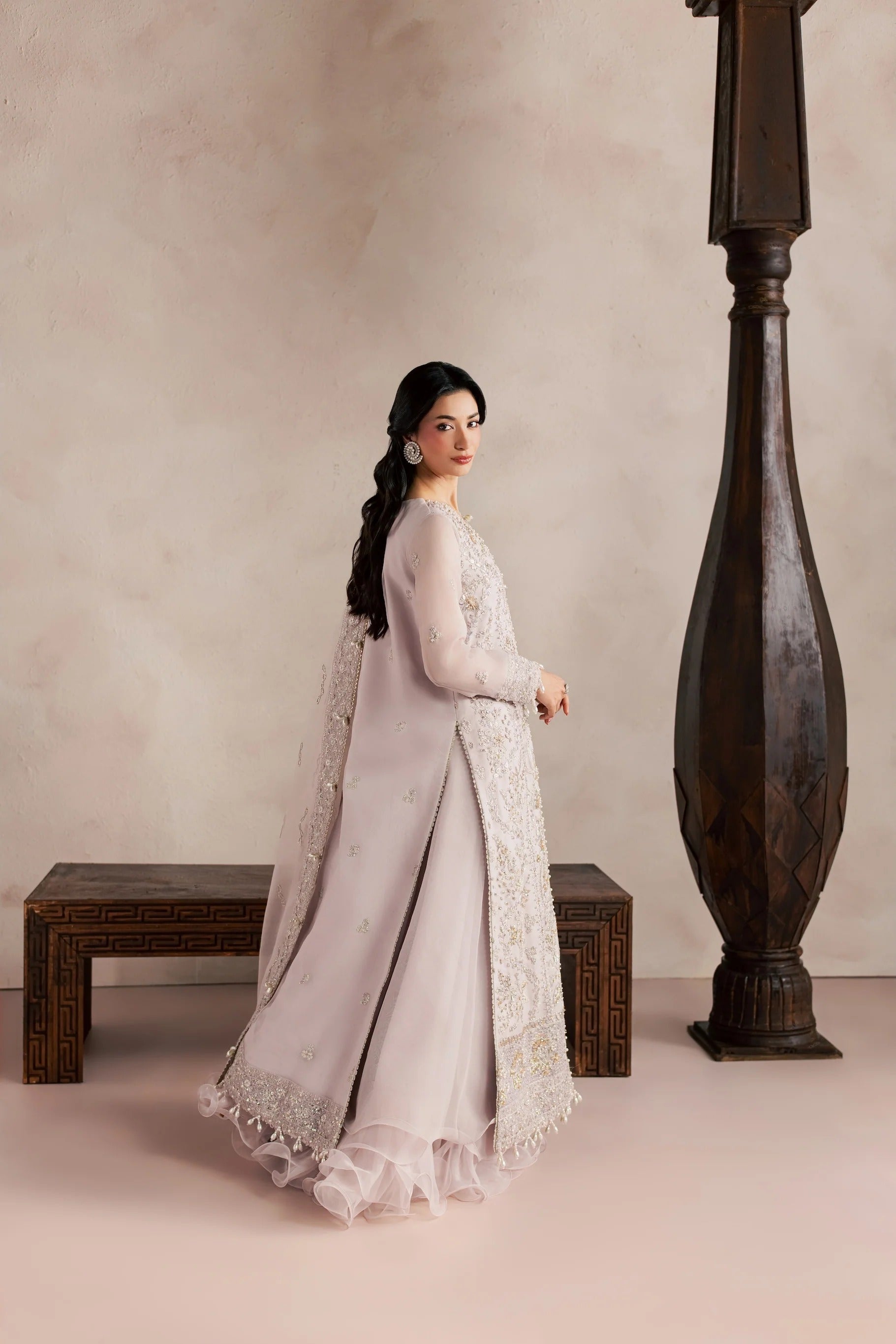 A woman wearing a light pink traditional outfit with intricate white embroidery, standing in a studio setting with a textured beige wall, a dark wooden bench, and a tall dark wooden pillar.