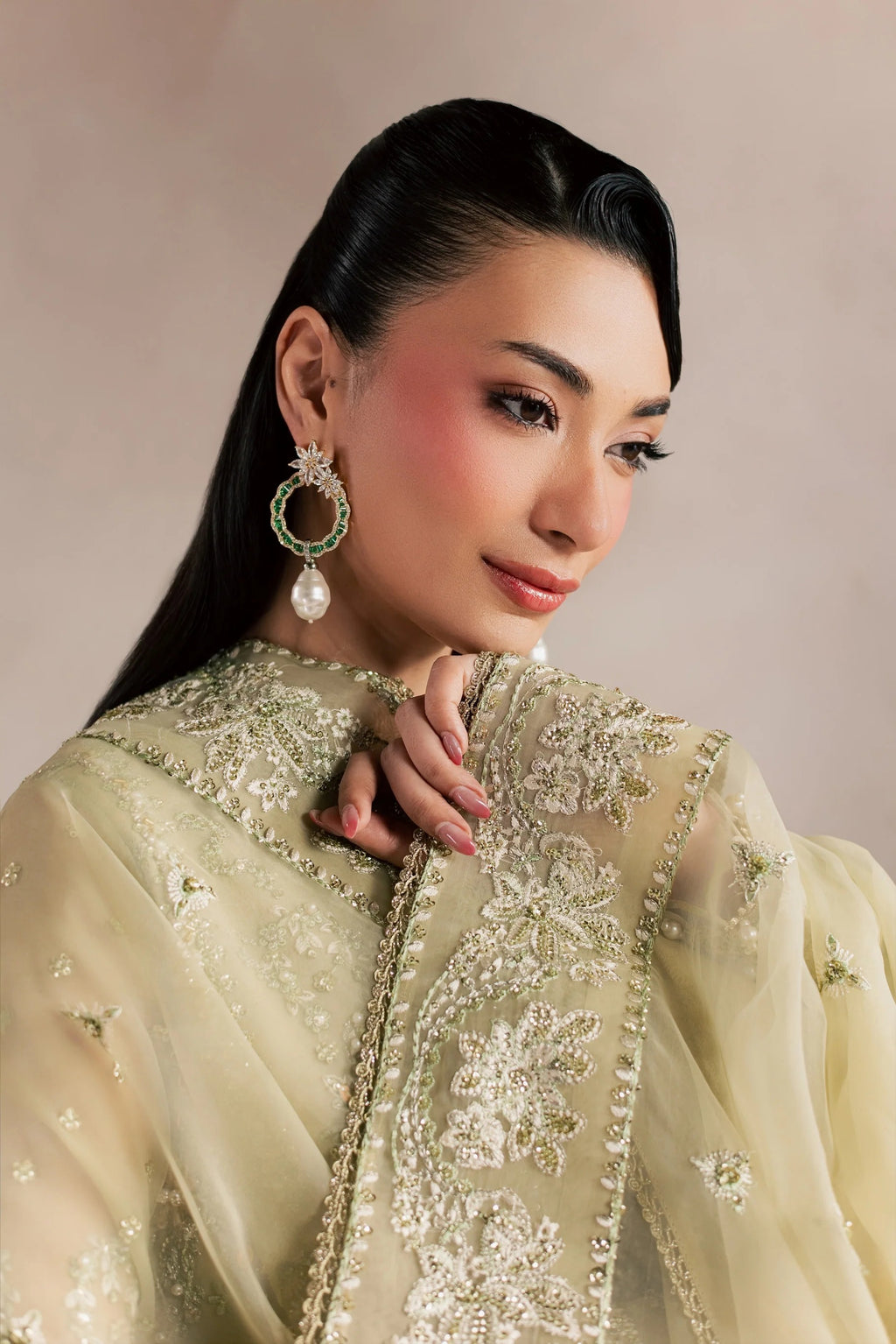 A woman wearing a traditional embroidered outfit in pale gold and beige with intricate metallic detailing, paired with a matching sheer draped scarf. She is wearing a large green and pearl earring, set against a soft beige studio background.