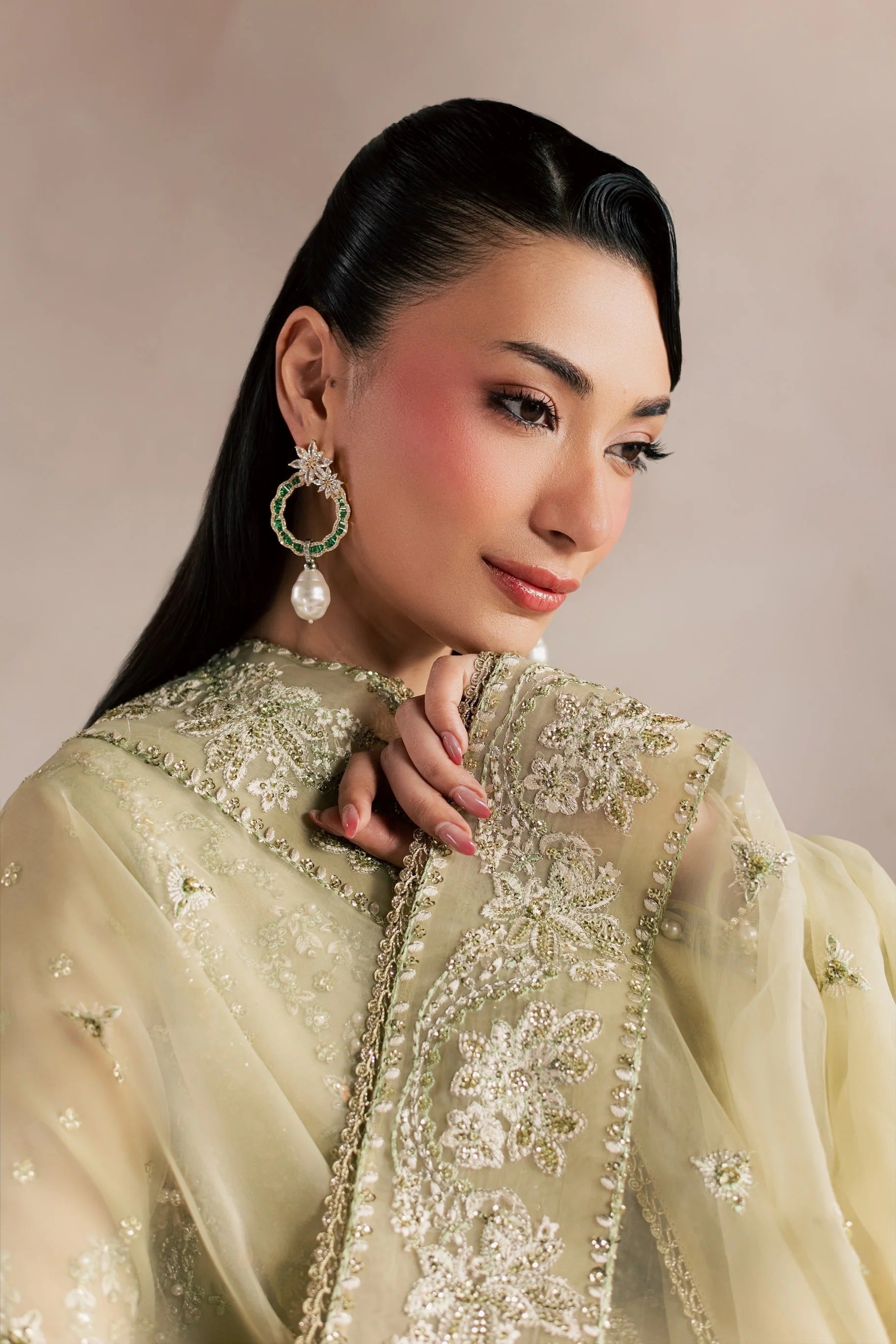 A woman wearing a traditional embroidered outfit in pale gold and beige with intricate metallic detailing, paired with a matching sheer draped scarf. She is wearing a large green and pearl earring, set against a soft beige studio background.