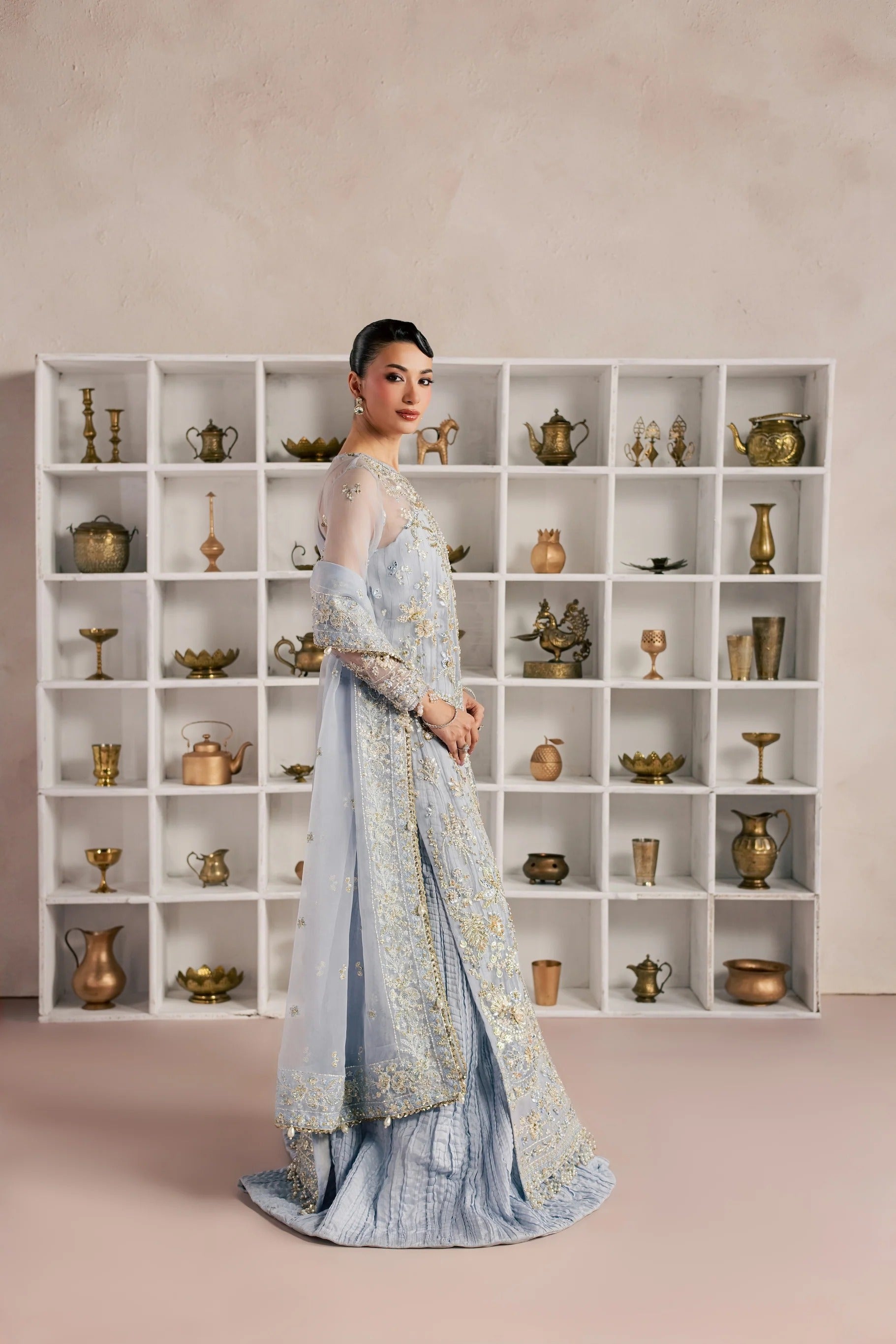 A woman models a light blue traditional outfit with intricate gold embroidery, standing in front of a white shelving unit filled with decorative brass and ceramic vessels against a textured beige wall.