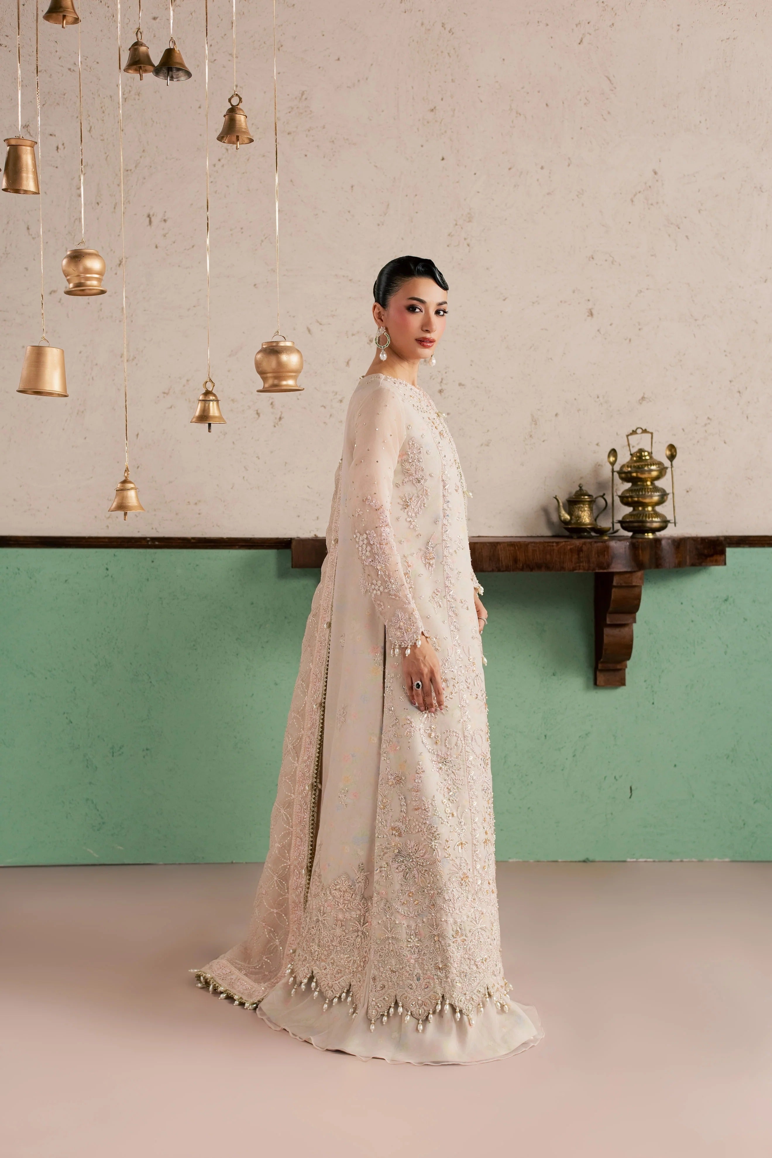 A woman models a long-sleeved cream-colored embroidered gown with a matching dupatta, standing in a styled room with a textured wall, hanging brass bells, and a wooden mantel.