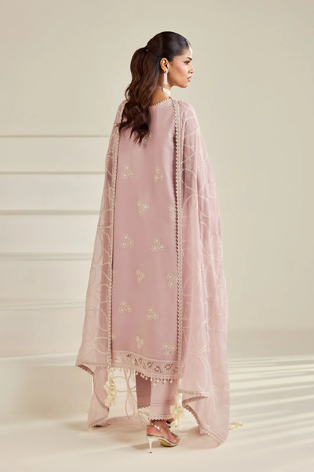 A woman modeling a dusty pink traditional outfit with a matching sheer dupatta, shown from the back against a warm beige background.