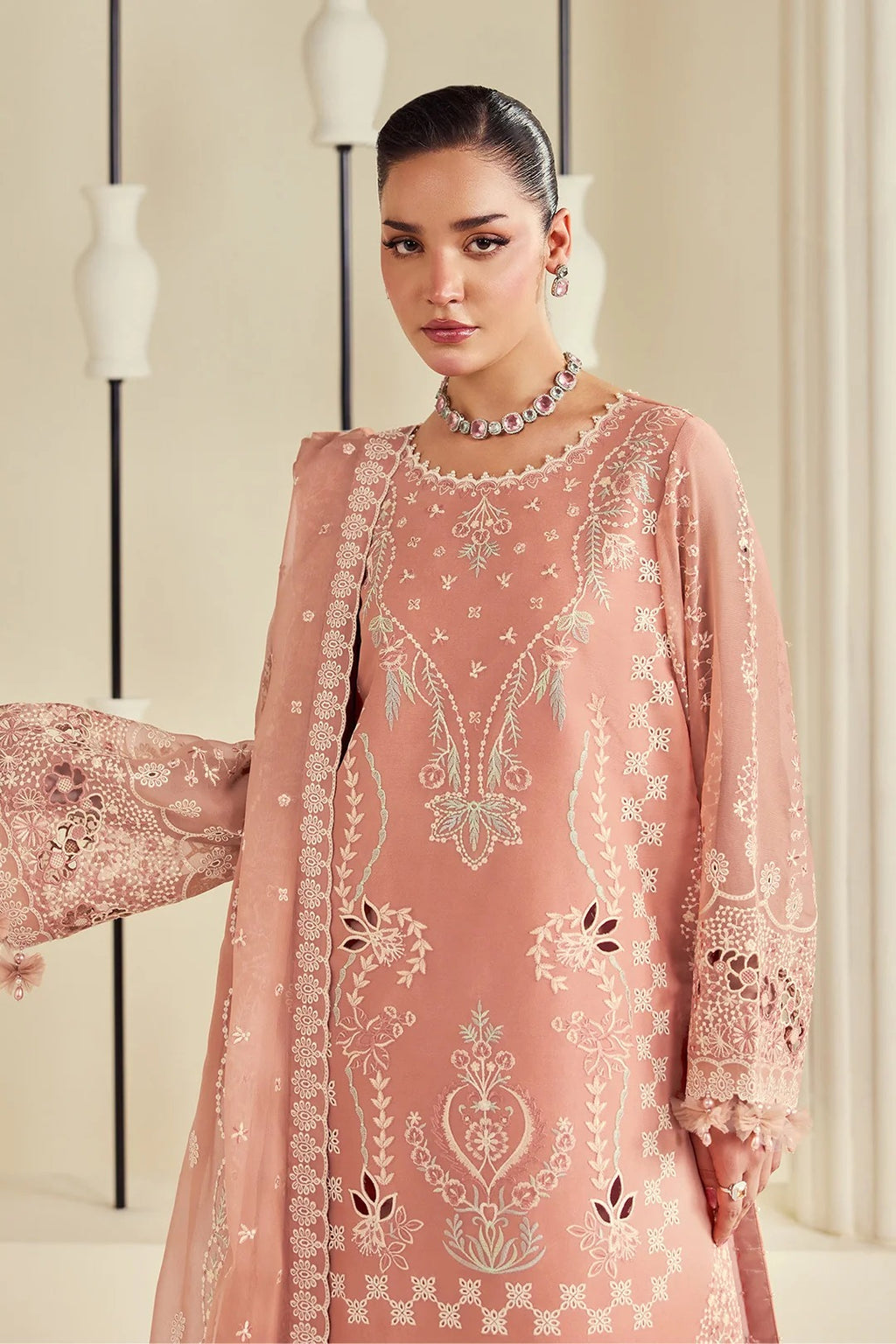 A woman modeling a peach-pink embroidered traditional outfit with a matching sheer dupatta, featuring intricate silver and white floral embroidery, set against a warm beige background with minimalist modern wall lamps.