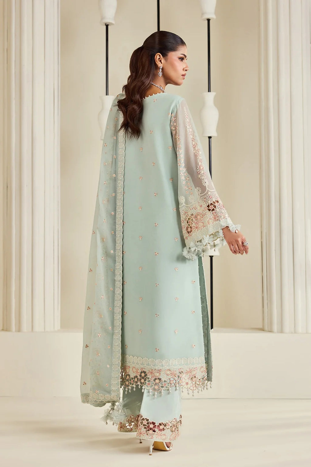 A woman modeling a mint green traditional outfit with floral embroidery and a matching dupatta, shown from the back in a studio setting with cream columns.