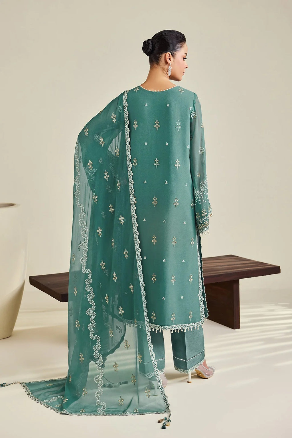 A woman stands with her back to the camera wearing a teal-green traditional outfit with matching dupatta and white embroidered motifs, photographed in a studio with a bench and neutral wall.