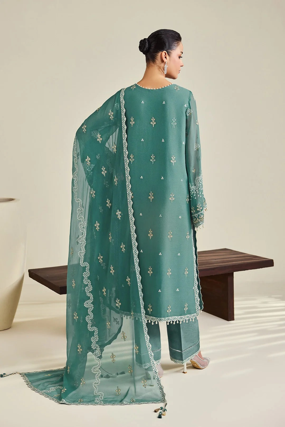 A woman stands with her back to the camera wearing a teal-green traditional outfit with matching dupatta and white embroidered motifs, photographed in a studio with a bench and neutral wall.
