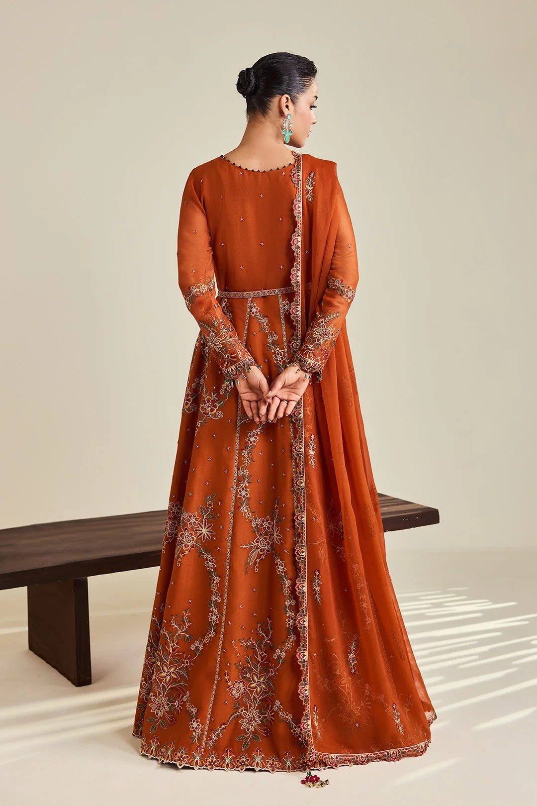 A woman stands with her back to the camera wearing a rust-orange traditional gown with intricate silver and gold embroidery, paired with a matching dupatta, in a studio setting with a wooden bench.
