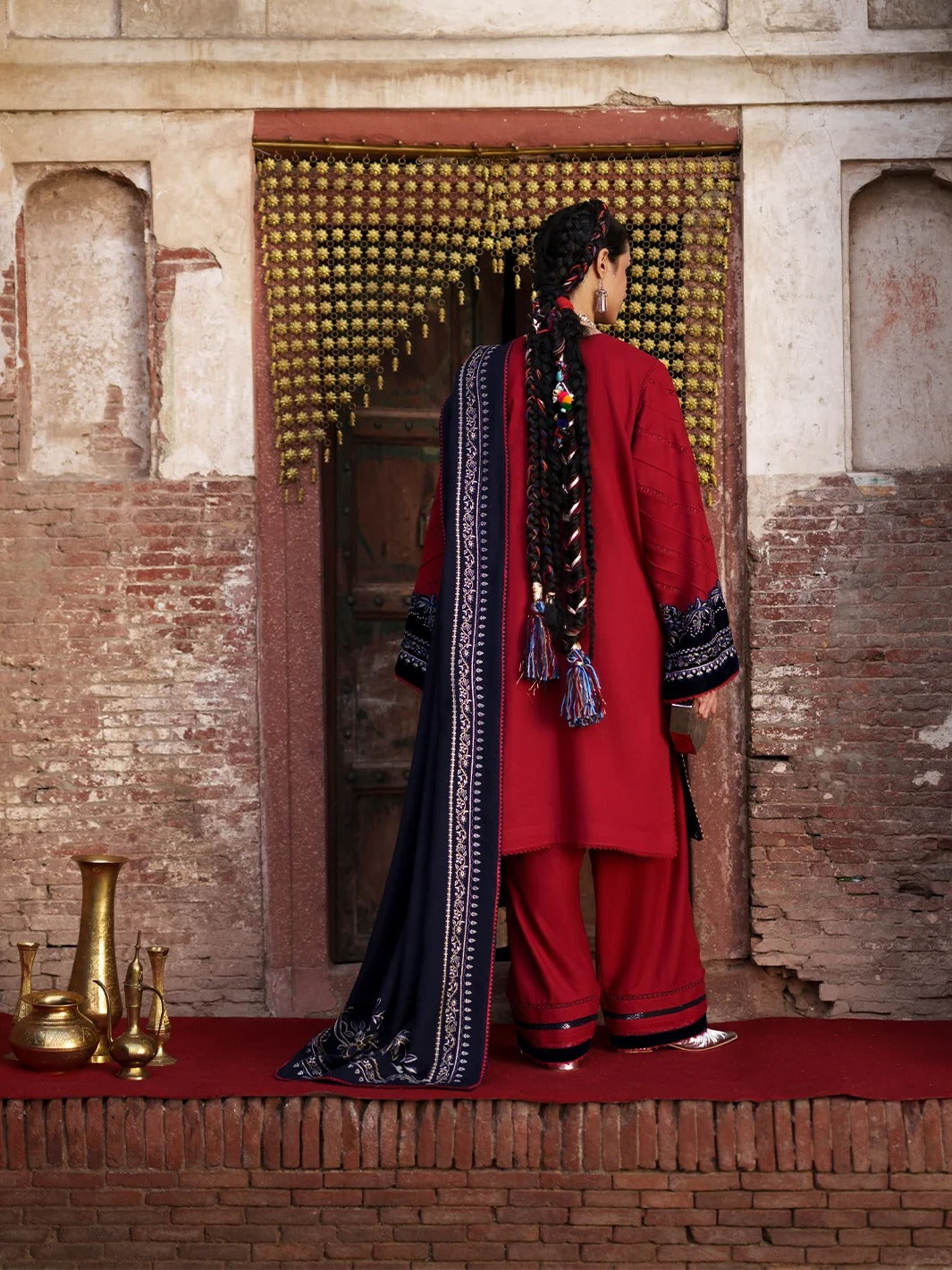 A woman stands with her back to the camera wearing a traditional red Indian outfit with a navy blue patterned dupatta, posed in front of a rustic brick doorway with brass hanging ornaments.